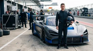 A man in a black racing suit holding a helmet stands next to a grey and blue race car in a pit lane