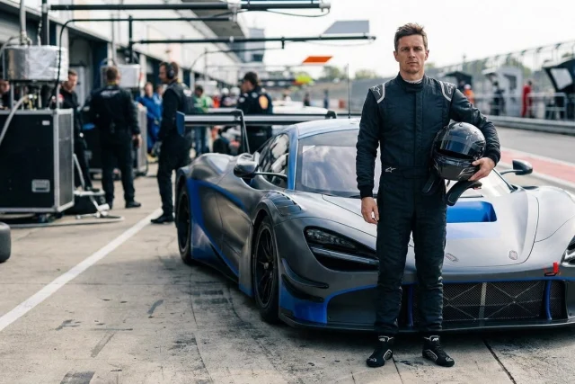 A man in a black racing suit holding a helmet stands next to a grey and blue race car in a pit lane