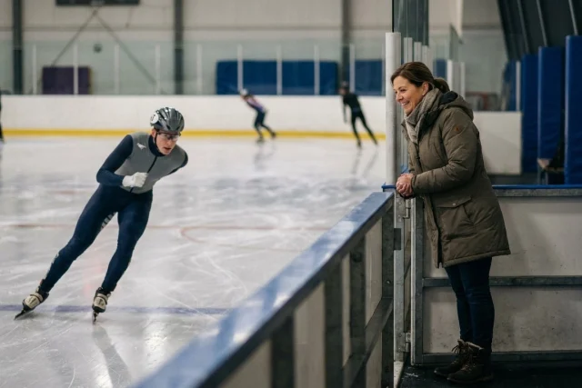 Woman in a winter coat stands smiling behind a barrier at an indoor ice rink, watching a speed skater in a grey and
